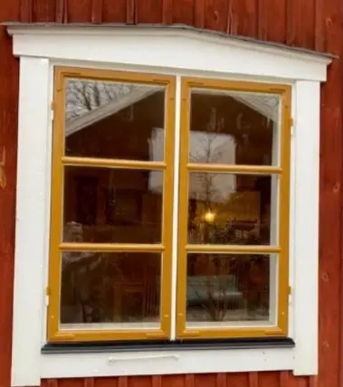 Ockramålat spröjsfönster med vitt foder på röd träfasad, efter renovering.