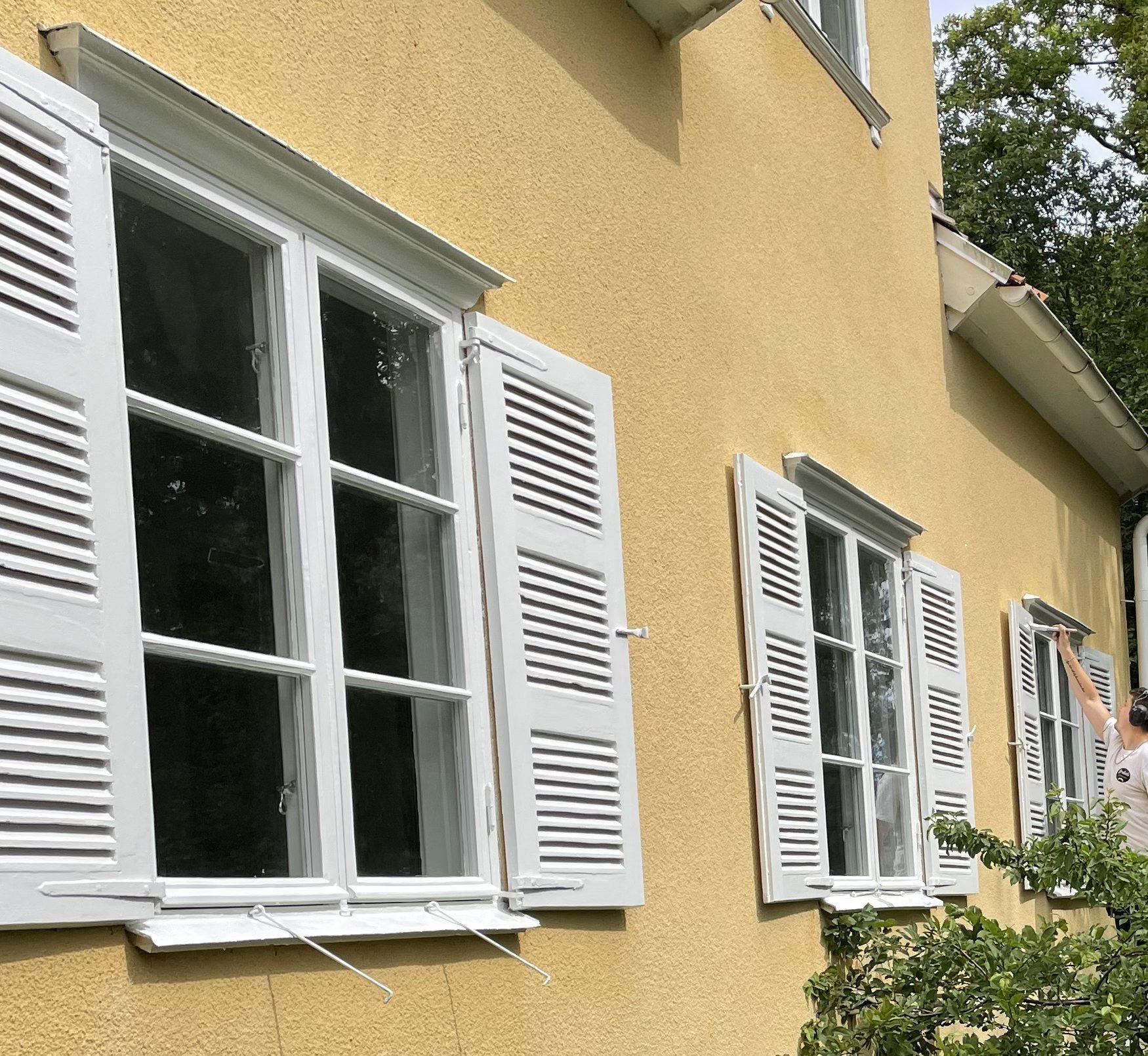 Gul putsad villafasad med vitmålade träfönster och fönsterluckor, sommargrönska omkring.