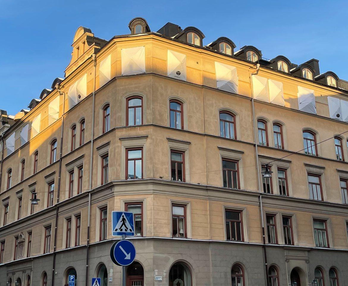 Fasad på sekelskiftesfastighet i Stockholm med rader av spröjsade träfönster i rödbruna karmar, i varmt kvällsljus.
